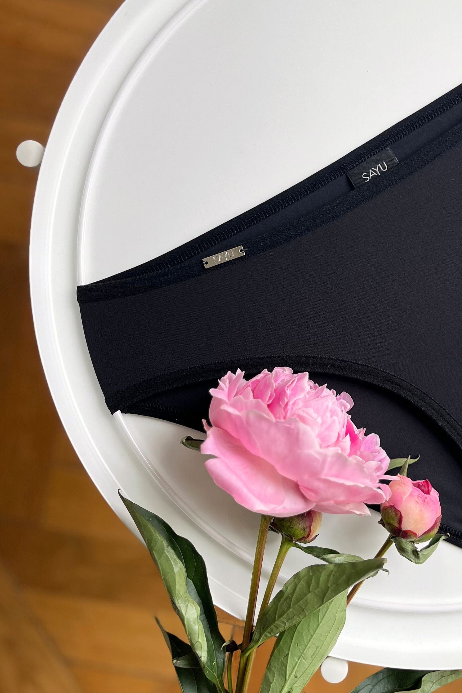 Detail na stříbrný zdobný SAYU prvek černých menstruačních plavek klasického střihu, pohled zepředu, foceno na bílém pozadí s květinou