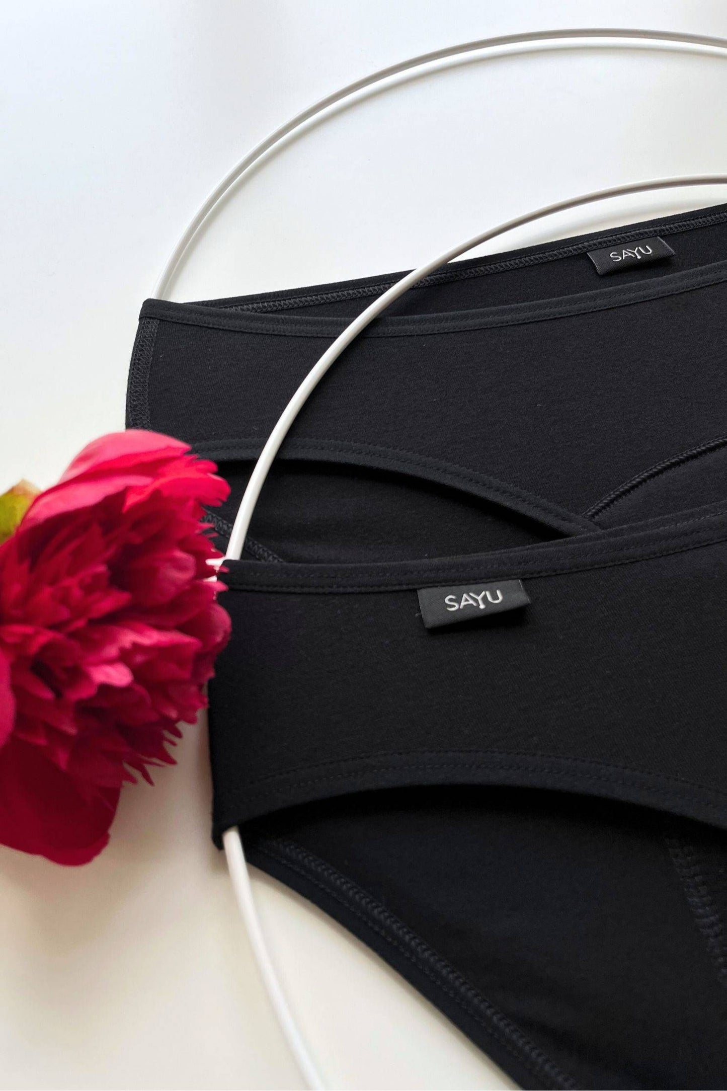Detail na zdobnou SAYU etiketu a švy menstruačních kalhotek na noc v černé barvě, pohled zepředu, foceno na bílém pozadí s květinou