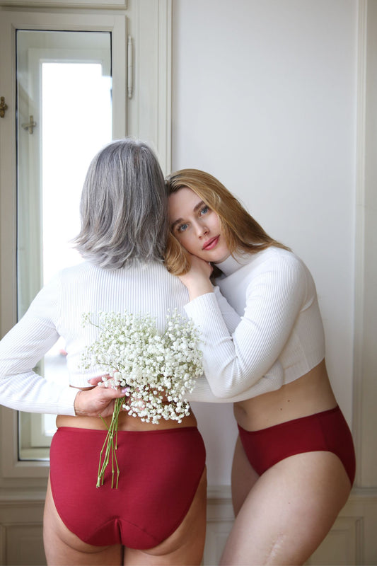 dvě modelky, které spolu stojí před oknem. Jedna drží v ruce bílou květinu s drobnými květy, obě na sobě mají menstruační merino kalhotky v bordó barvě
