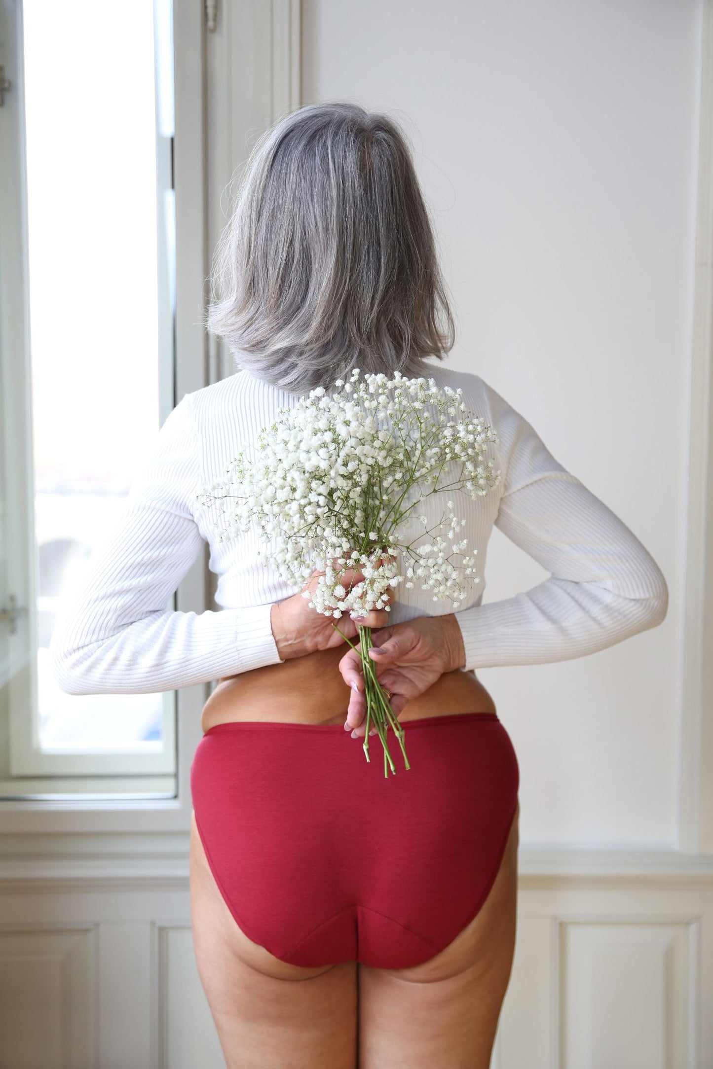 pohled zezadu na modelku, která v rukou drží bílé květiny s drobnými kvítky - na sobě má bordó merino kalhotky klasického střihu