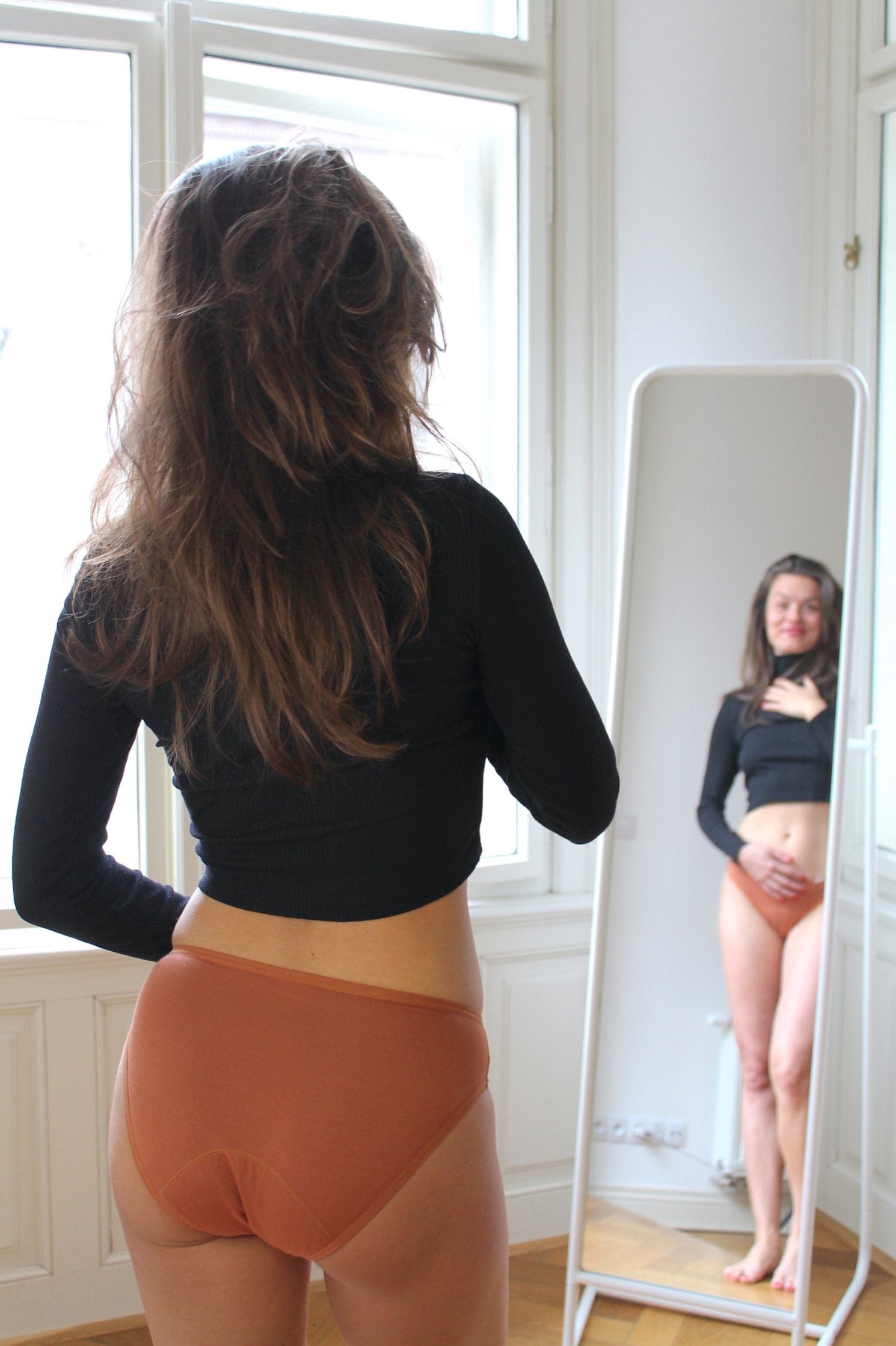 pohled na modelku, která se na sebe kouká do zrcadla a má na sobě menstruační kalhotky SAYU klasické skořicové v nízké savosti