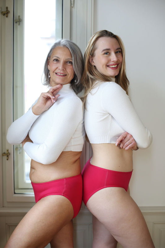 dvě modelky stojící k sobě blízko zády, usmívající se do objektivu - na sobě mají klasicky střižené menstruační kalhotky ve svěže růžové barvě