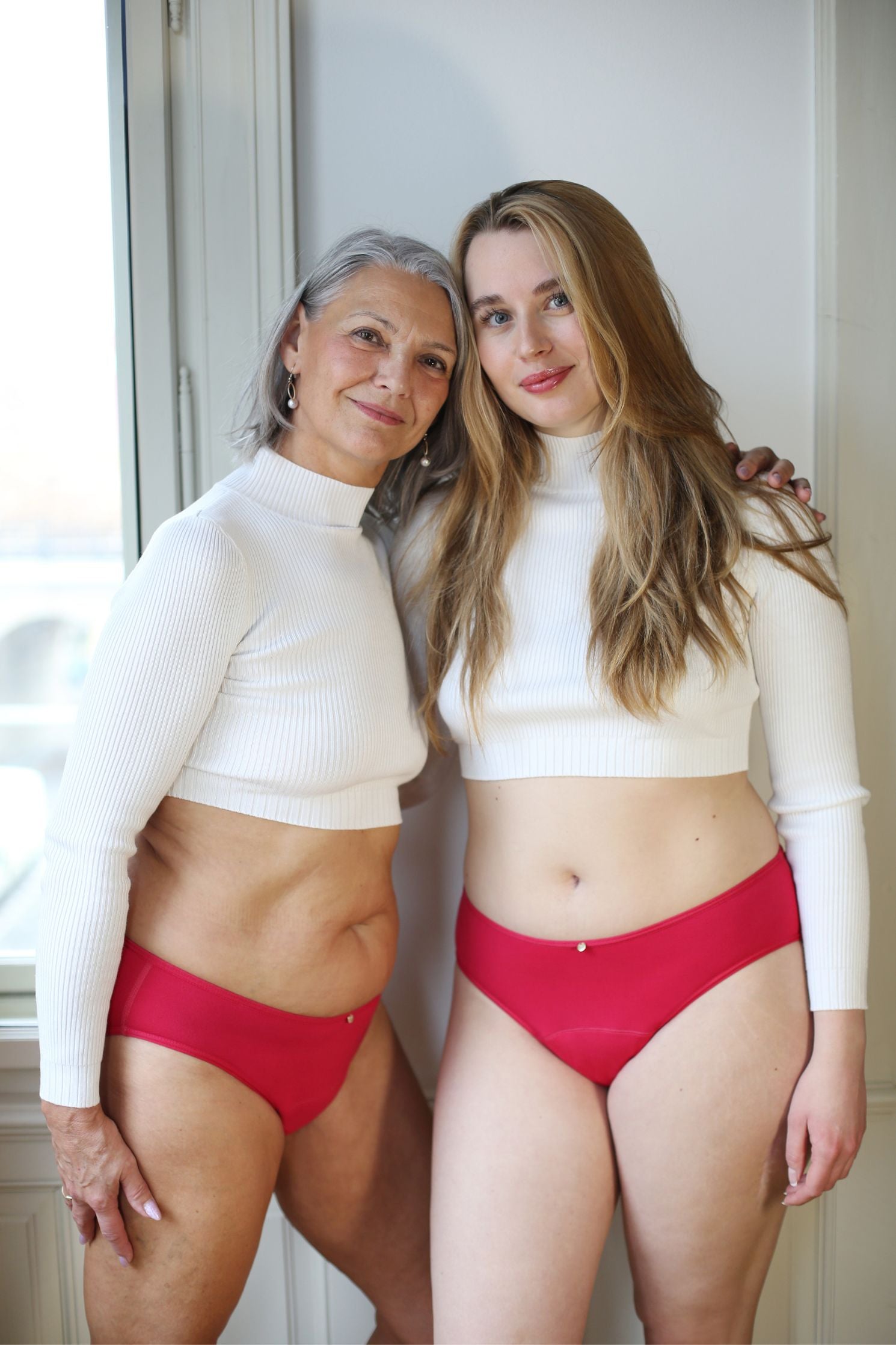 dvě modelky stojící před oknem, usmívající se do objektivu - na sobě mají klasicky střižené menstruační kalhotky ve svěže růžové barvě