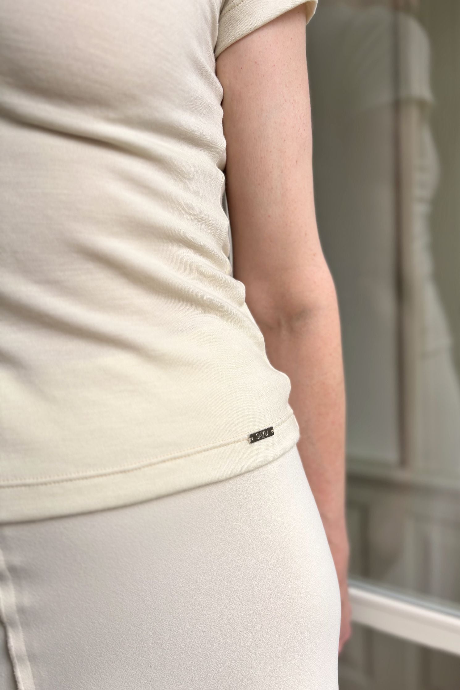 Detailní pohled na zdobný prvek na SAYU merinovém tričku v přírodní barvě.